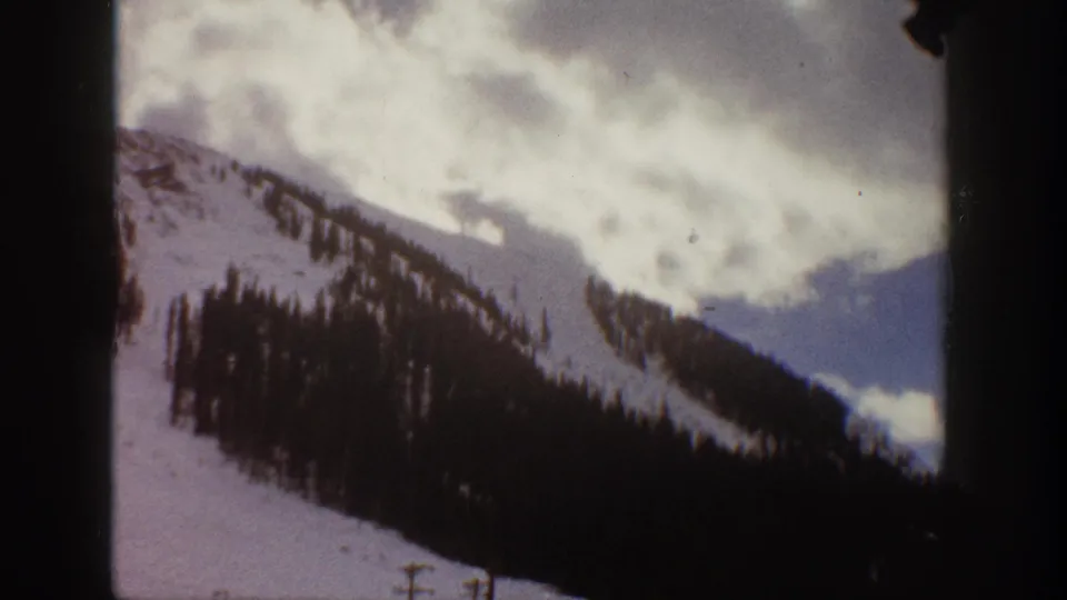 1961 Aspen-area frame showing a snow-covered ridge, dark conifers, clouds, and distant blue mountains.