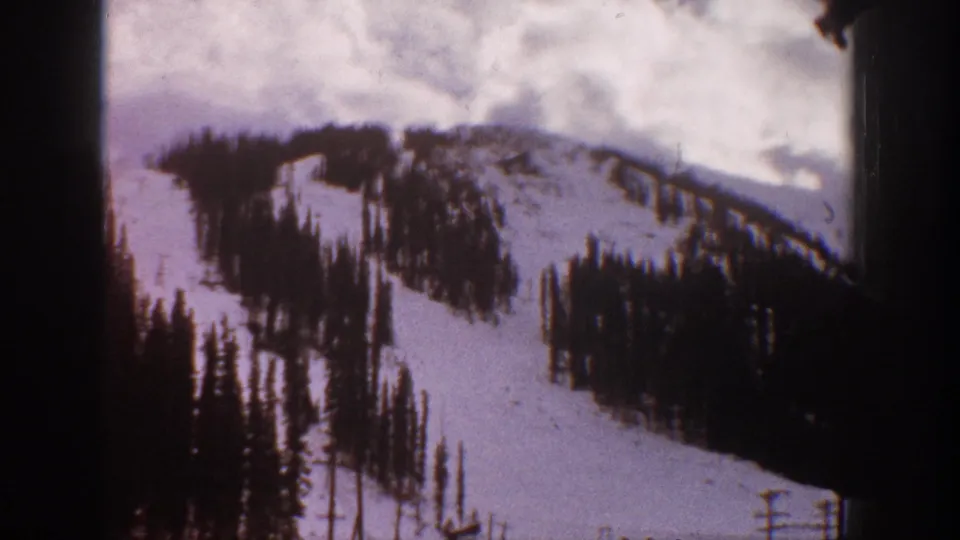 1961 Aspen, Colorado frame showing a steep snowy slope with tall evergreen trees and a cloudy winter sky.