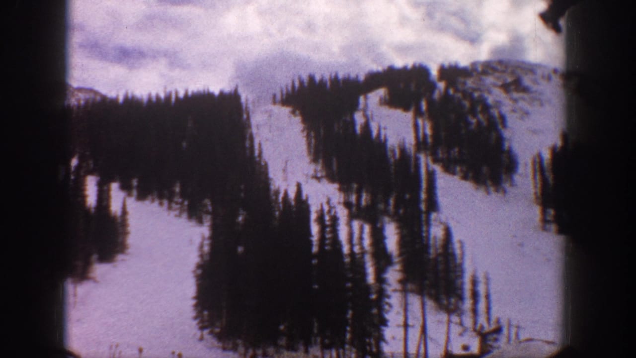 Snowy ski slope with evergreen trees and mountain ridges, seen through film-frame edges.