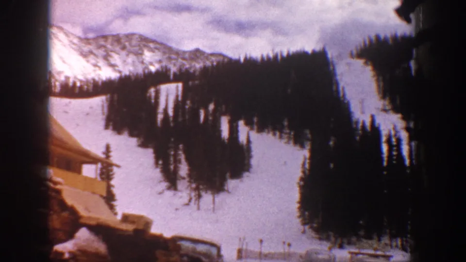1961 Aspen home-movie frame showing a chalet-like lodge roof beneath snowy ski runs and dark evergreen slopes.