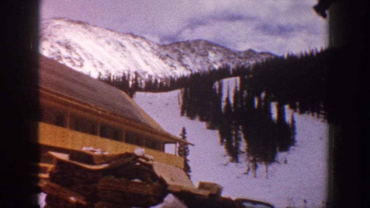 1961 Aspen home-movie frame showing a wooden building below snow-covered mountain slopes and dark evergreen trees.