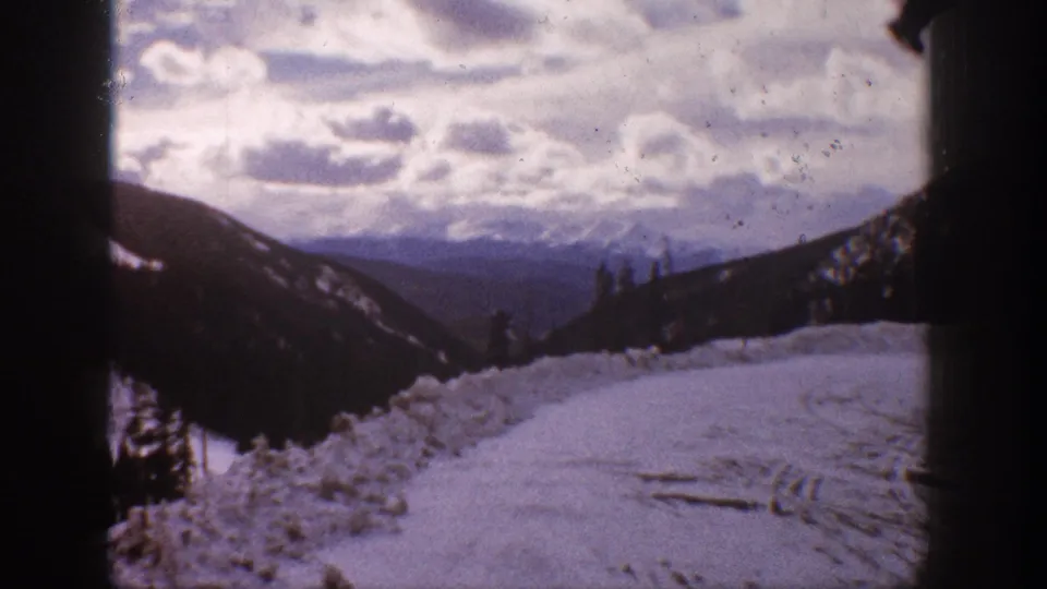 1961 Aspen winter view from a snowy road, looking past dark slopes toward distant mountains and clouds.