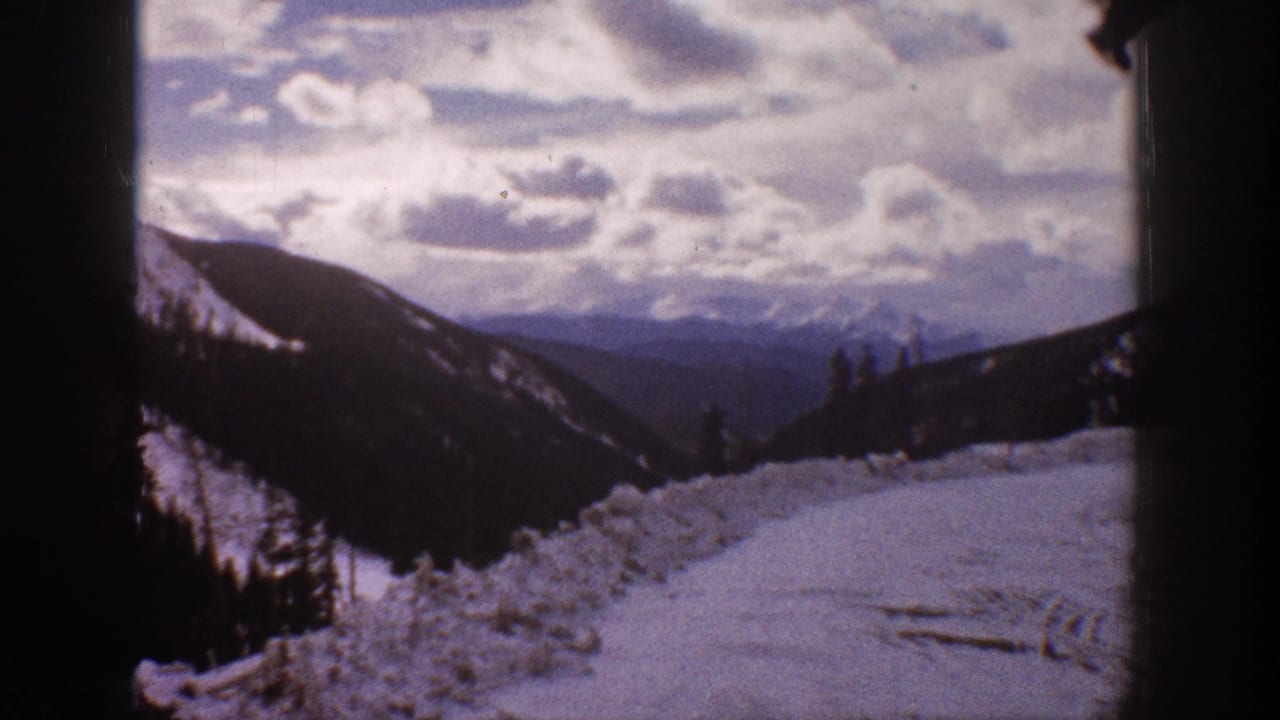1961 Aspen, Colorado home-movie frame showing a snow-covered mountain road or slope, forested ridges, and a cloudy winter sky.
