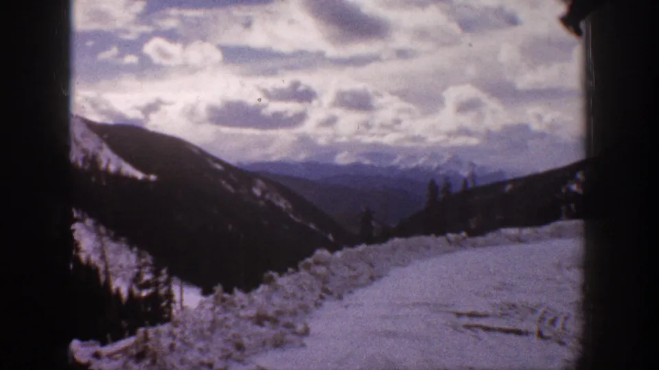 1961 Aspen, Colorado home-movie frame showing a snow-covered mountain road or slope, forested ridges, and a cloudy winter sky.