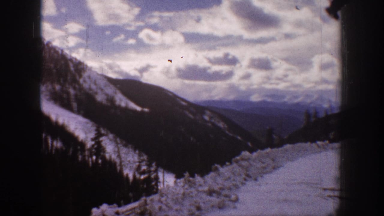 Snowy mountain ridge and valley with evergreen trees beneath a bright clouded sky.