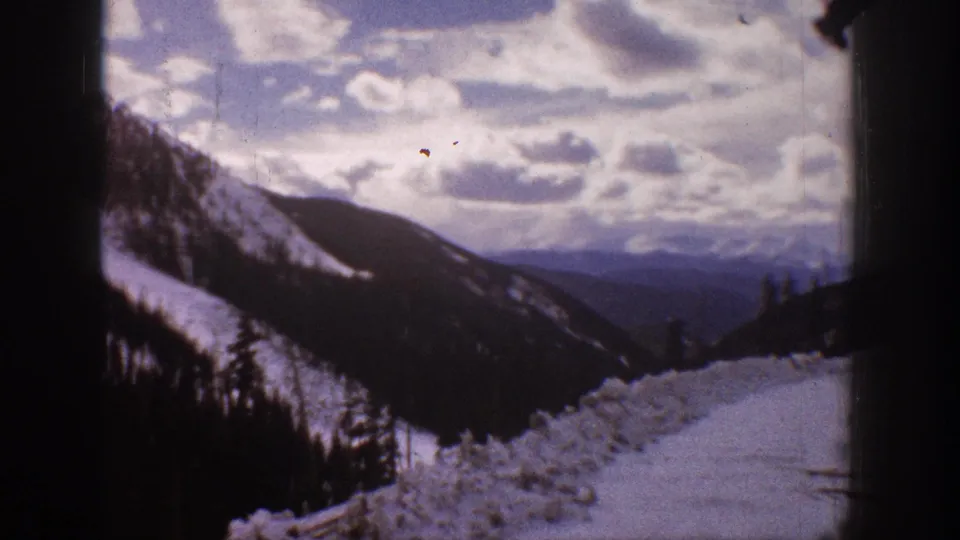 1961 Aspen, Colorado frame with a snow-covered ridge, dark evergreen forest, distant mountains, and bright clouds.
