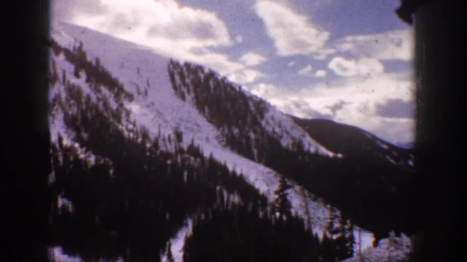 1961 Aspen, Colorado frame showing a broad snow-covered slope, evergreen trees, distant ridges, and cloudy sky.