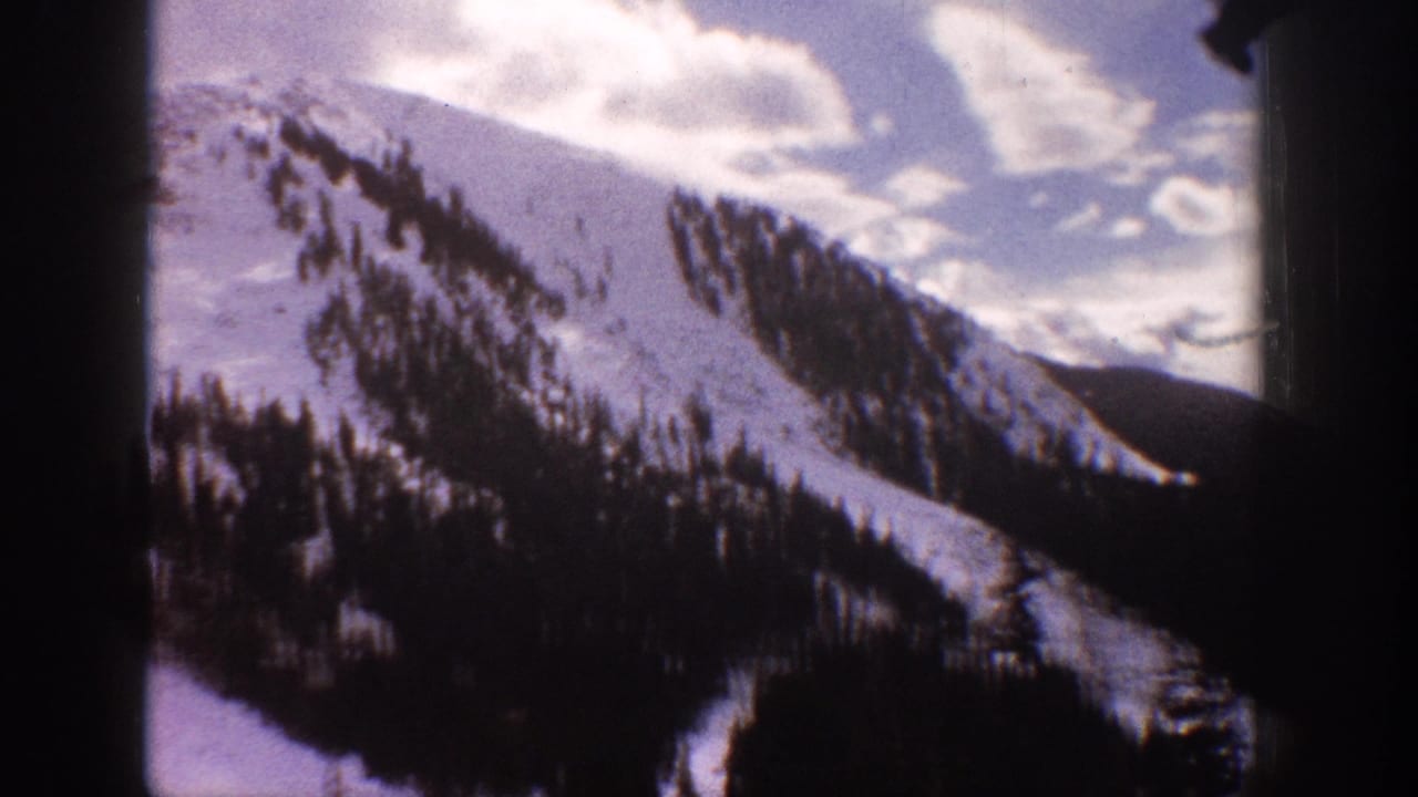 Snowy mountain slope with evergreen stands under a cloudy sky, framed by dark film borders.