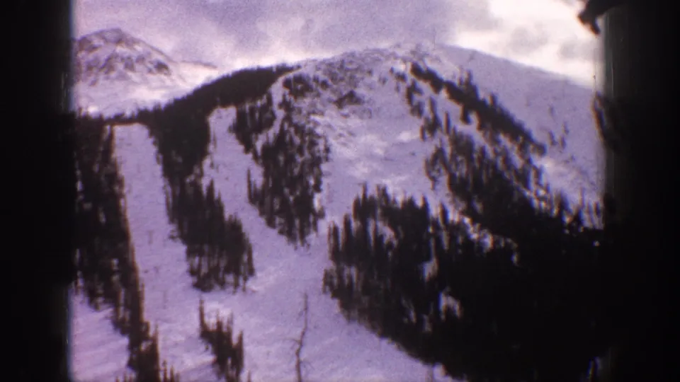 1961 Aspen, Colorado 8mm frame showing a wider snowy slope, dark evergreen bands, distant ridge forms, and soft home-movie color.