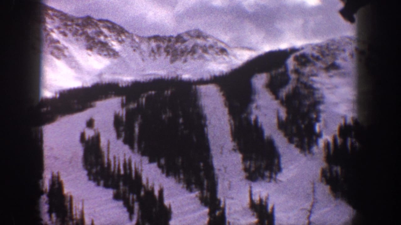 1961 Aspen, Colorado home-movie frame showing snow-covered slopes, dark evergreen stands, and curving ski trails beneath a cloudy sky.