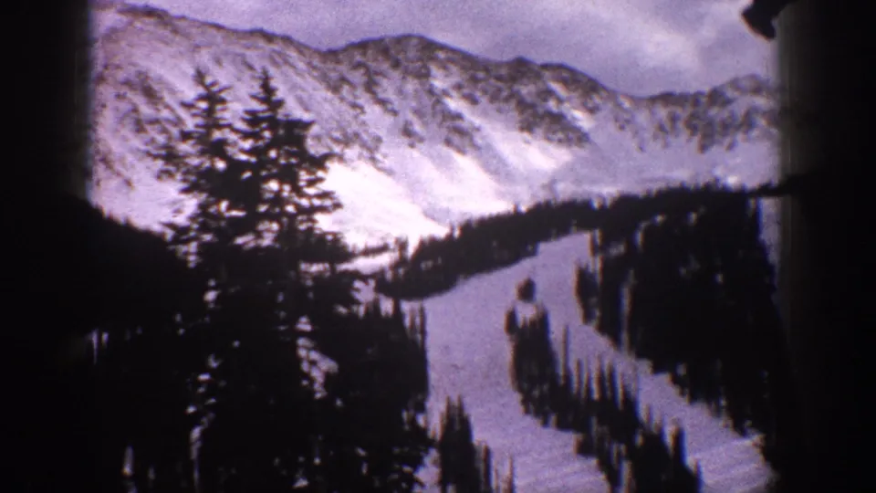 1961 Aspen home-movie view with foreground evergreens, snowy ski terrain, and a pale mountain ridge softened by 8mm film grain.