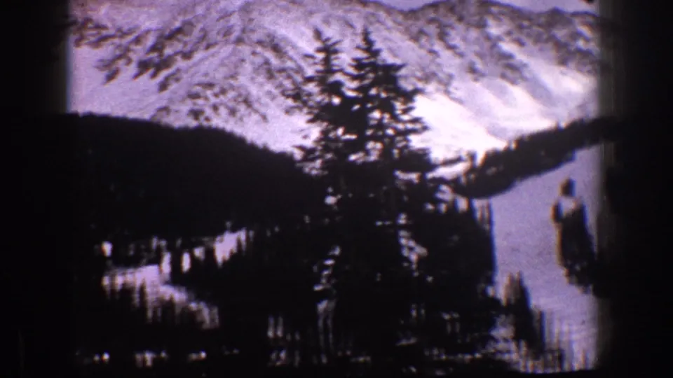 Aspen, Colorado 1961 8mm frame showing a snow-covered mountain face, clustered evergreens, and a curving ski run with dark foreground shadow.