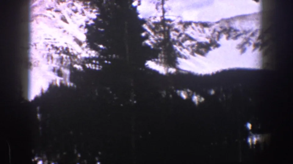 Vintage 8mm view of a snow-covered Aspen mountain face seen through dark foreground evergreens.