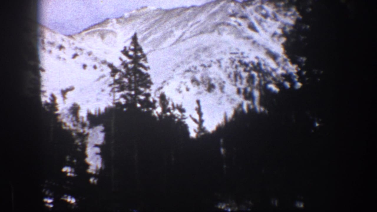 1961 Aspen, Colorado home-movie frame showing a snow-covered mountain ridge behind a dark evergreen foreground.
