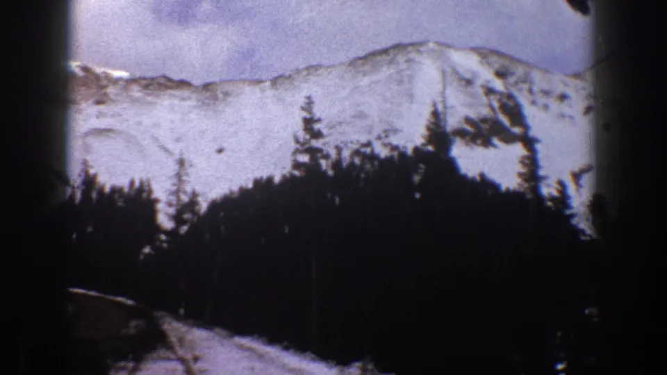 1961 Aspen, Colorado frame showing a snowy ridge, dense evergreens, and a small roofline or structure at the lower edge.