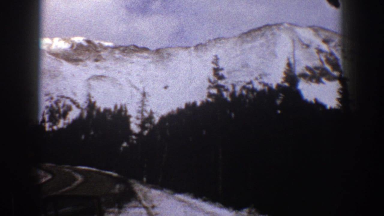 1961 Aspen, Colorado home-movie frame showing a winding snowy road from inside a car, with dark windshield edges, trees, and mountain slopes.