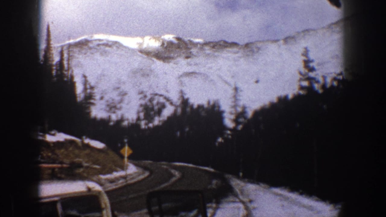 1961 Aspen, Colorado frame from inside a car, looking toward a curve in the snowy mountain road with a small roadside sign visible.