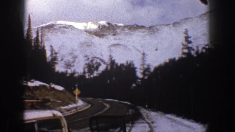1961 Aspen, Colorado frame from inside a car, looking toward a curve in the snowy mountain road with a small roadside sign visible.