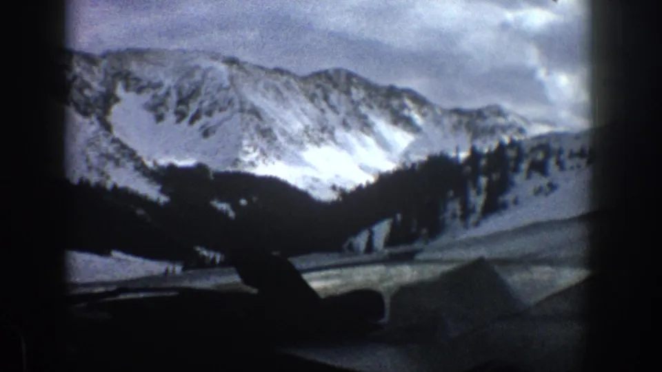 1961 Aspen, Colorado home-movie view across a car dashboard toward snow-bright mountain slopes and dark evergreen ridges.