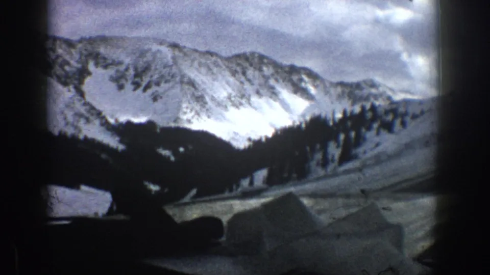 1961 Aspen, Colorado frame showing the car’s dashboard and windshield view toward snowy alpine slopes, trees, and a road edge.