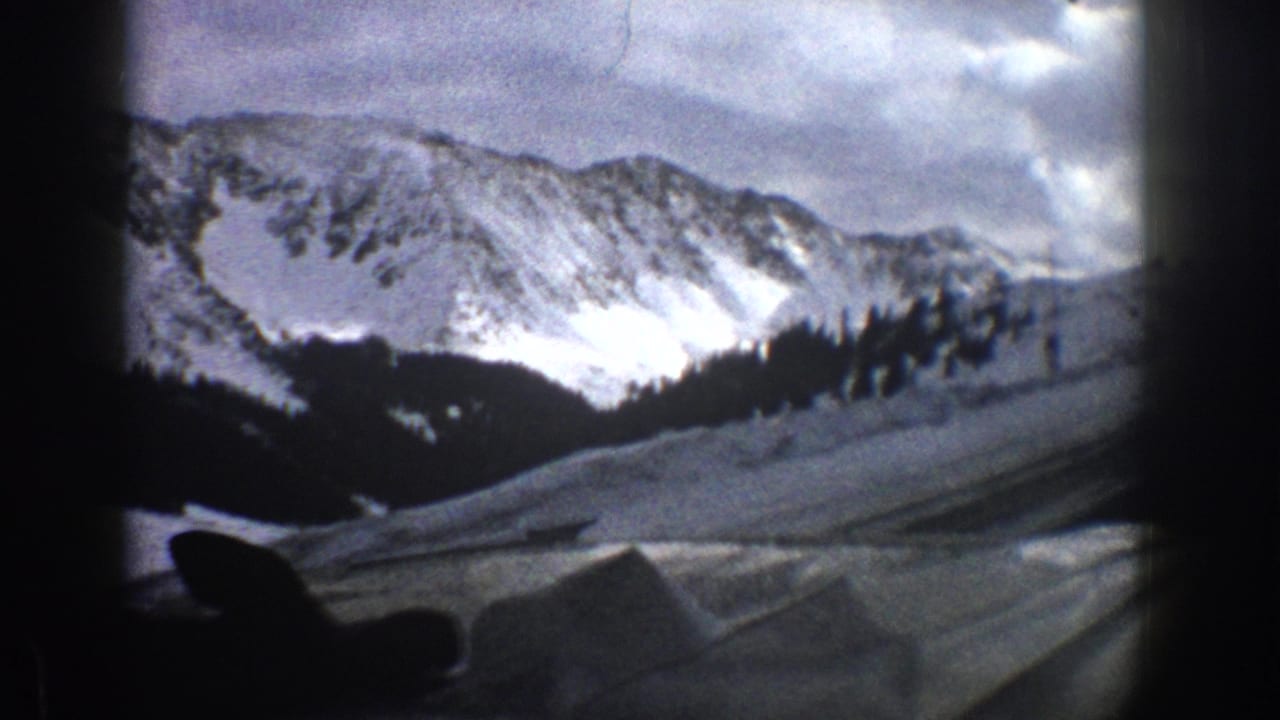 1961 Aspen-area 8mm frame showing snowy mountain peaks, evergreen slopes, and a vehicle-side foreground under cloudy sky.