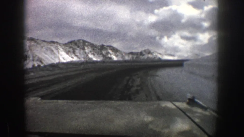 Aspen, Colorado 1961 travel frame showing a snow-lined mountain road view, distant ridge, cloudy sky, and vehicle foreground detail.