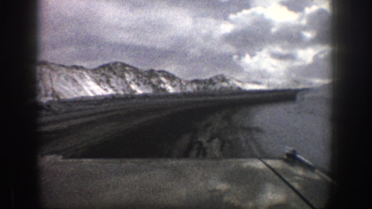 Dashboard view of a dark mountain road beside snowy ridges under a cloudy sky.