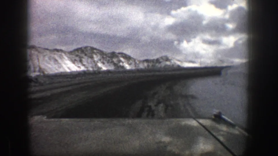 1961 Aspen, Colorado 8mm frame showing a windshield view of a winding road, snow-covered mountains, and heavy clouds.