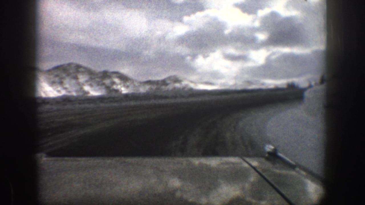 1961 Aspen, Colorado road-view frame with the dashboard at bottom, snowy ridges ahead, and high clouds over the mountains.