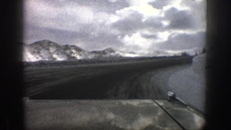 1961 Aspen, Colorado road-view frame with the dashboard at bottom, snowy ridges ahead, and high clouds over the mountains.