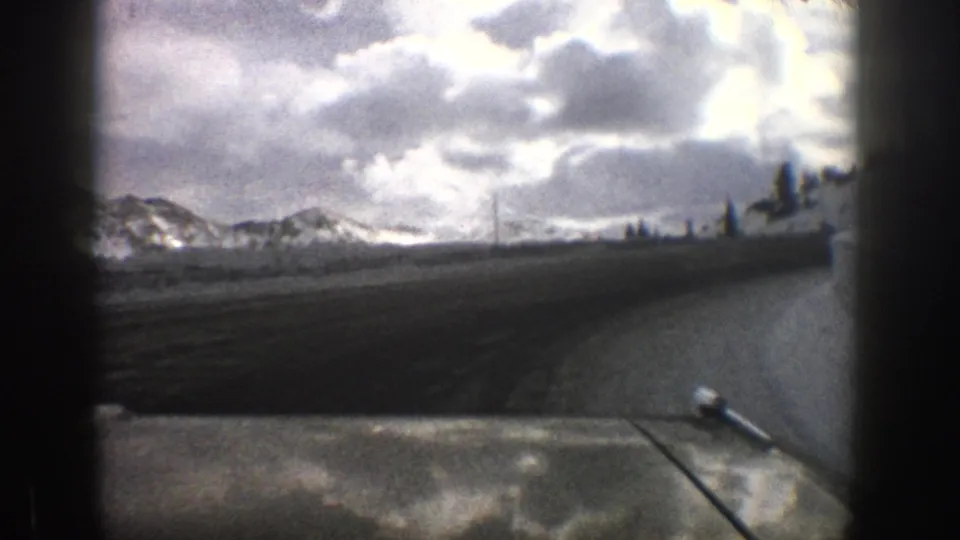 1961 Aspen, Colorado frame from inside a car showing a dark road, snow-covered terrain, distant mountains, and poles near the roadside.