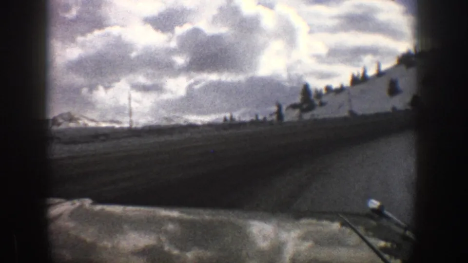 1961 Aspen, Colorado 8mm frame showing a windshield view past the dashboard toward a snowy hillside and mountain road under clouds.