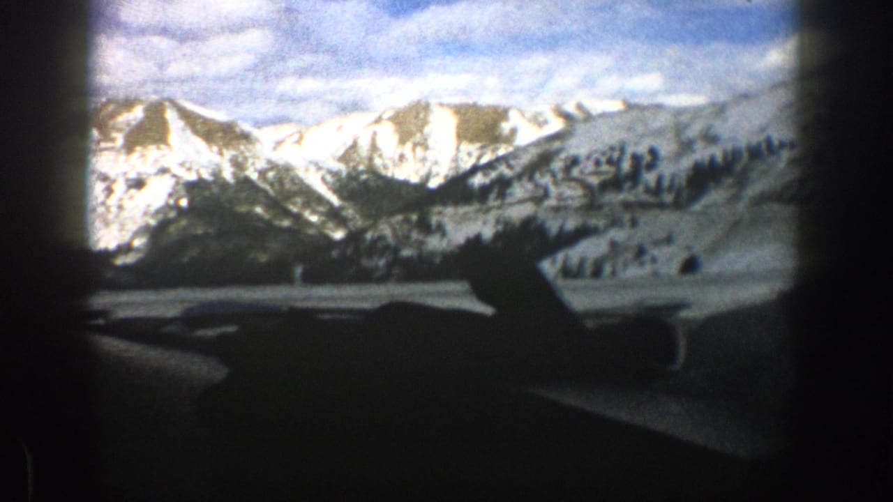 1961 Aspen winter mountain view from inside a moving car, with snowy peaks and dashboard silhouette visible.