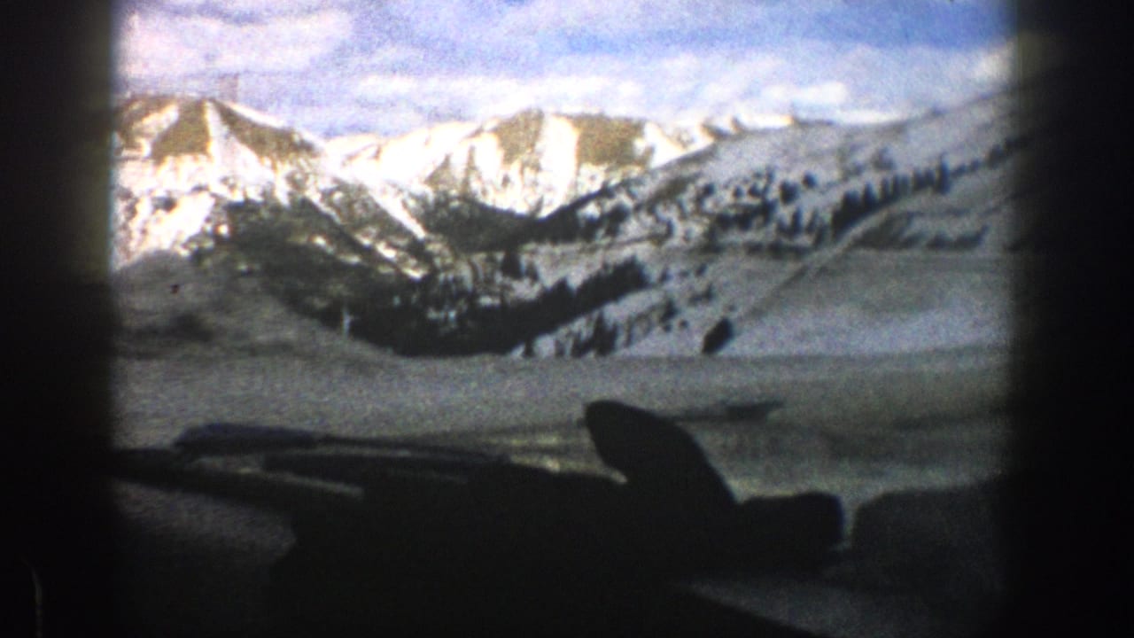 1961 Aspen-area home-movie view of snow-covered mountains through a vehicle windshield, with dashboard and wiper silhouettes in front.