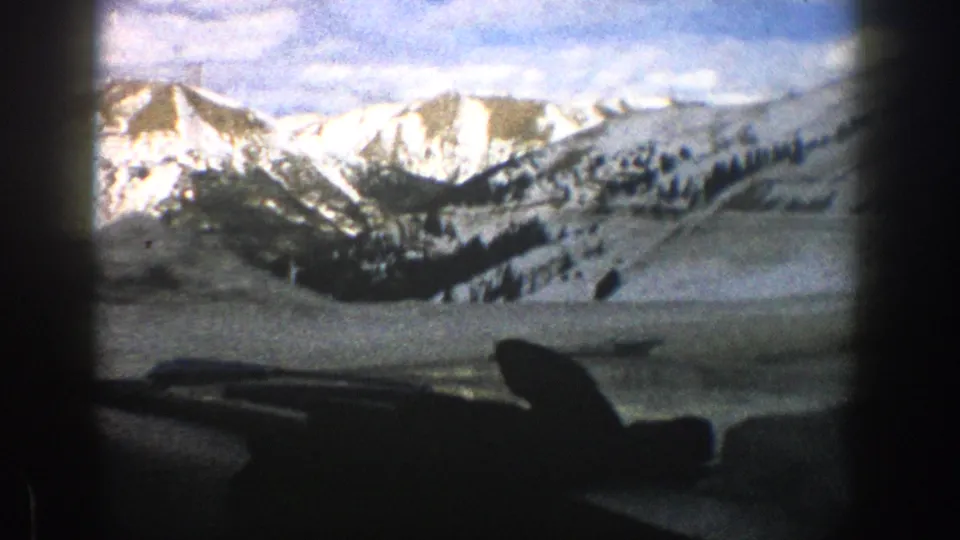 1961 Aspen-area home-movie view of snow-covered mountains through a vehicle windshield, with dashboard and wiper silhouettes in front.