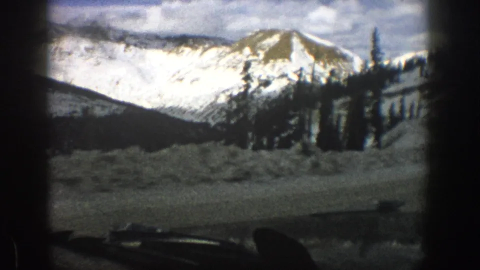 Final windshield view of a 1961 Colorado mountain pass, showing a bright snow-covered slope, trees, and dark vehicle interior shapes.