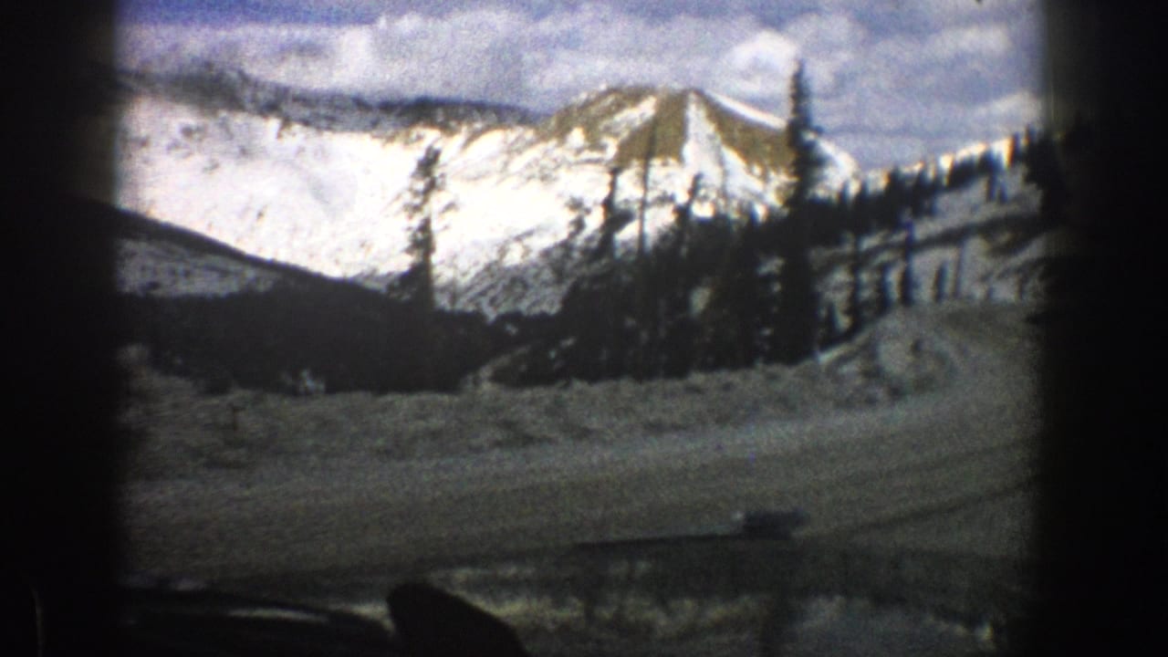 1961 Aspen-area 8mm view from inside a moving car toward a snowy road, dark evergreens, and sunlit mountain peaks.