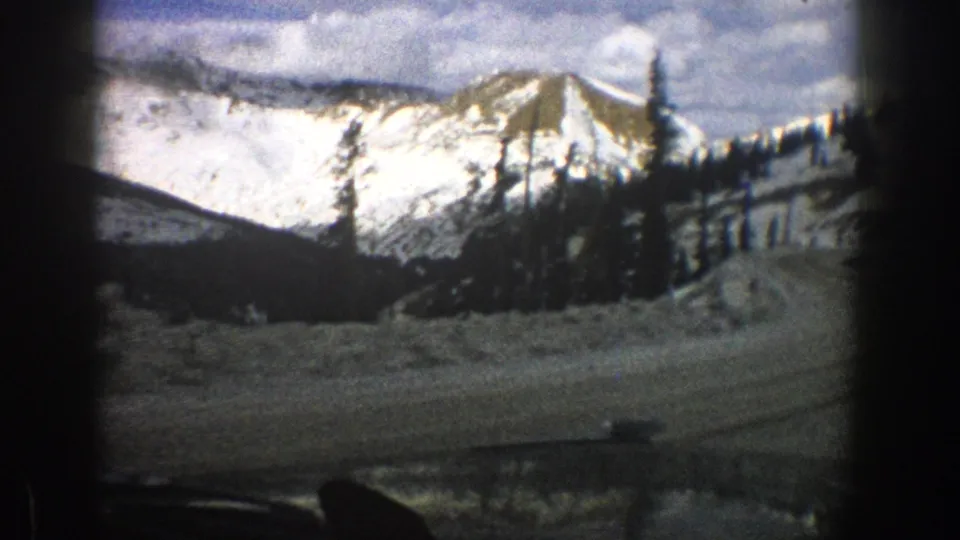 1961 Aspen-area 8mm view from inside a moving car toward a snowy road, dark evergreens, and sunlit mountain peaks.