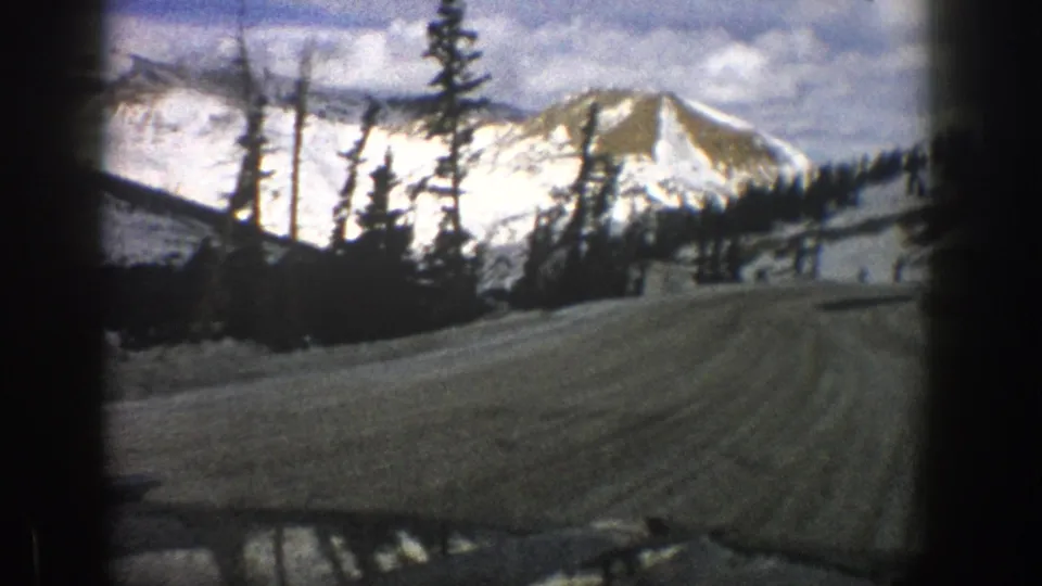 1961 8mm passenger-view frame near Aspen showing a snow-lined mountain road, evergreen silhouettes, and a bright ridge.