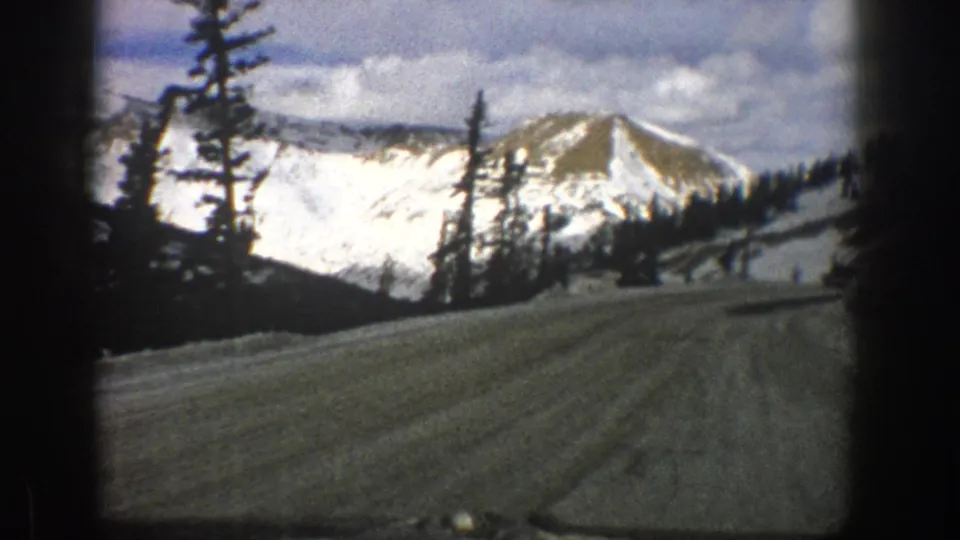 Aspen-area 1961 8mm frame from a vehicle on a snowy dirt road, with tall evergreens and white mountain slopes ahead.