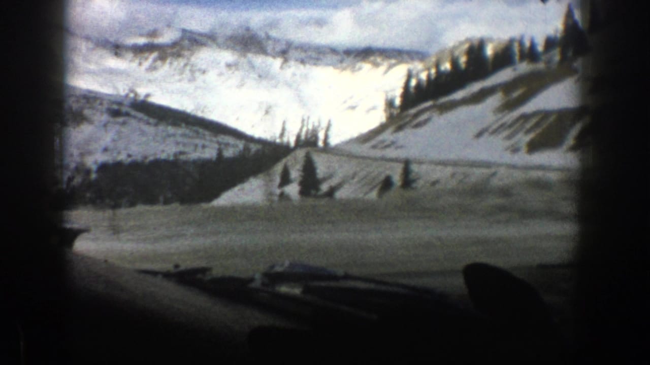 1961 Aspen-area 8mm frame showing snow-covered mountain slopes, scattered evergreens, cloudy sky, and the dark edge of a car interior.