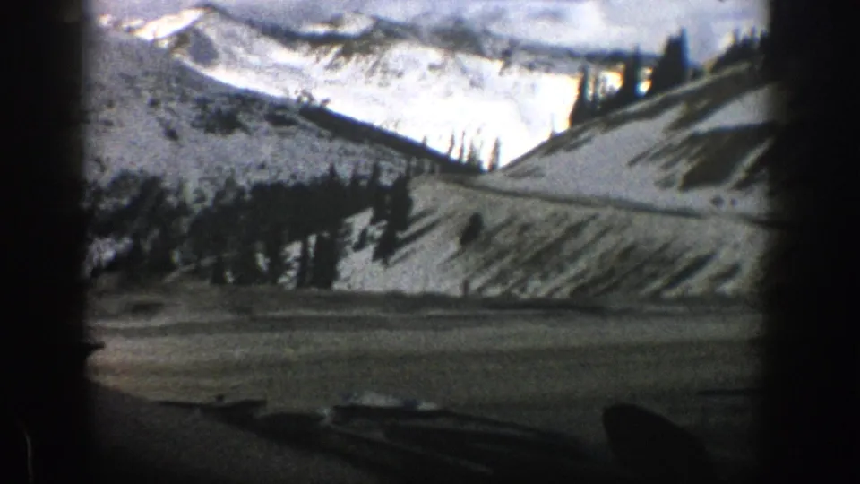 8mm home-movie frame from a car near Aspen showing a snowy roadside slope, clustered evergreens, and pale mountain ridges beyond.