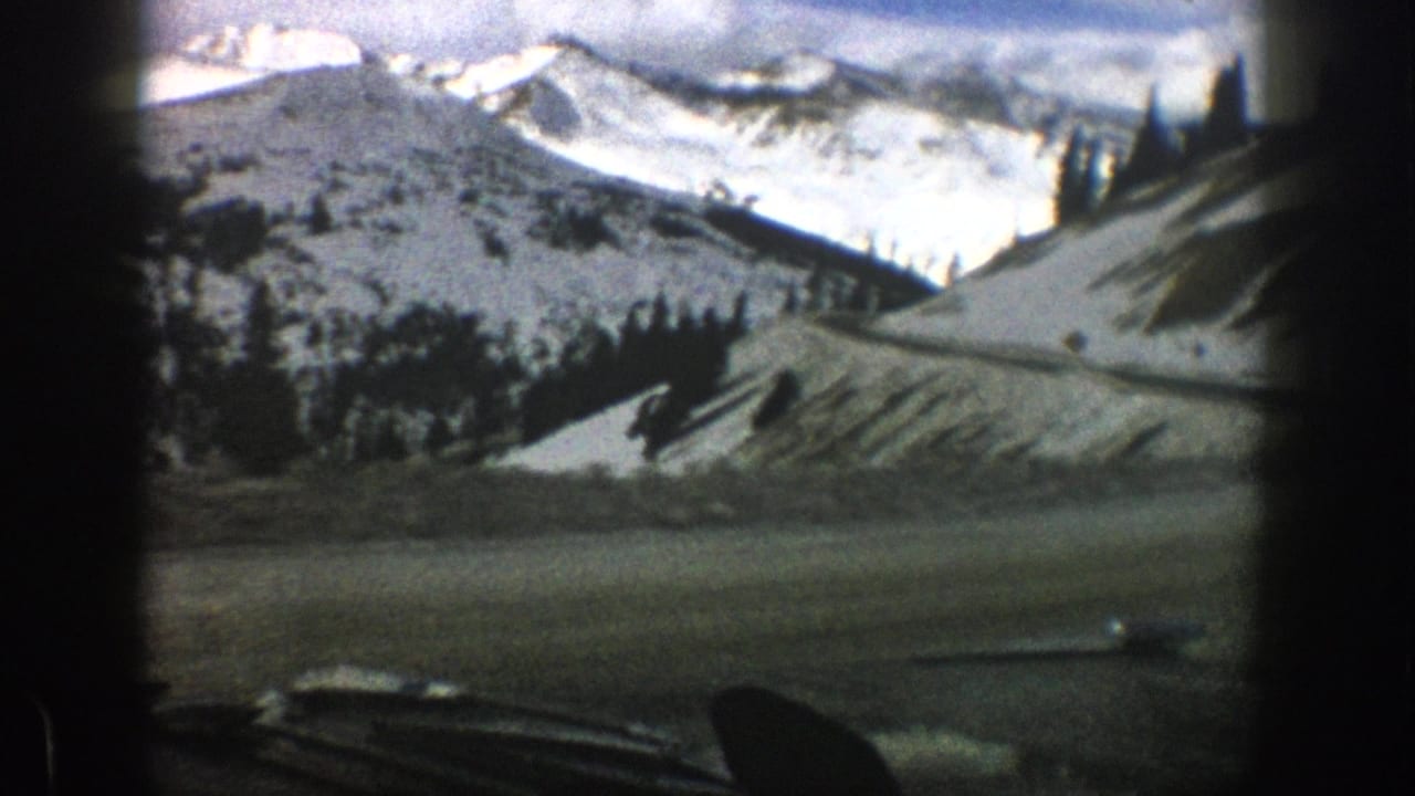 1961 Aspen, Colorado 8mm frame showing snow-dusted mountain slopes, scattered evergreen trees, cloudy sky, and dark foreground framing from a vehicle viewpoint.