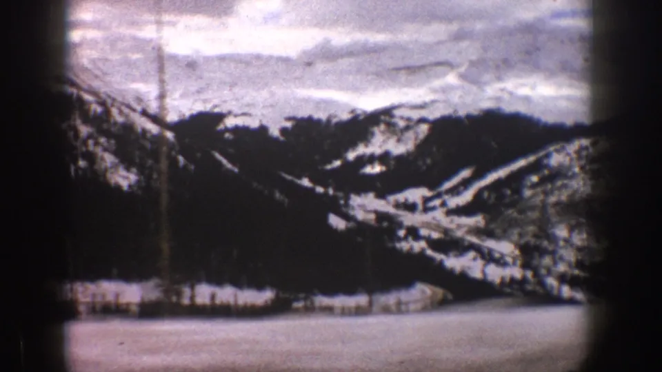 1961 Aspen, Colorado 8mm frame of a snowy foreground, dark evergreen ridge, distant mountain slopes, cloudy sky, and aged-film vignette around the scene.