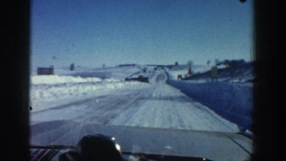 1961 Aspen-area road view from a moving vehicle, with a snowy lane, roadside banks, blue sky, and distant low structures or posts.