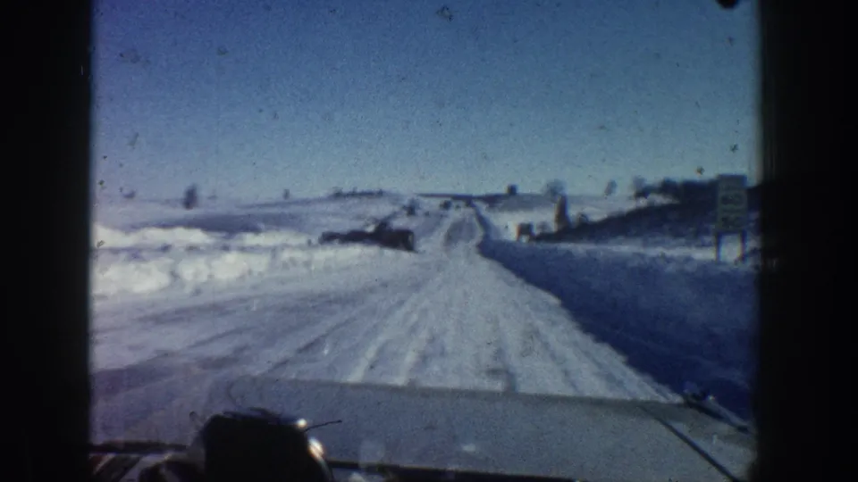 1961 Aspen-area 8mm frame showing a moving vehicle perspective on a snow-packed road under clear blue sky, with roadside snowbanks and distant objects.