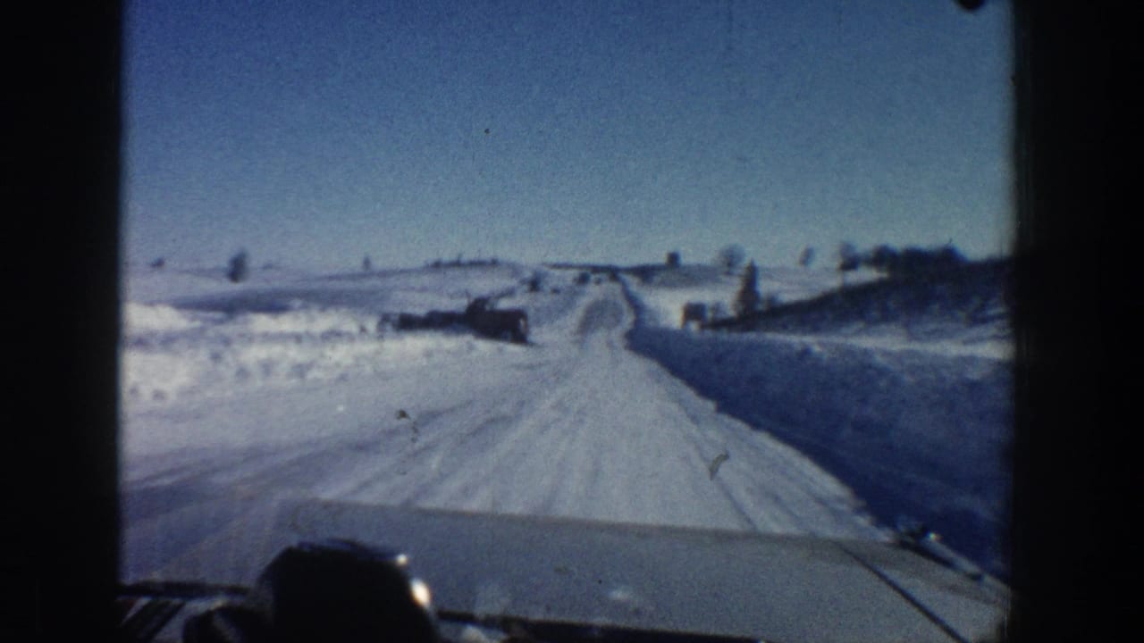 Snow-covered rural road seen over a car hood, with snowbanks and distant buildings under blue sky.