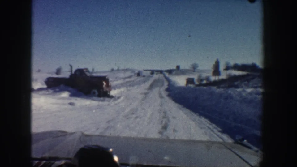 1961 Aspen-area home-movie frame with a car passing a dark truck along a narrow snow-covered rural road.