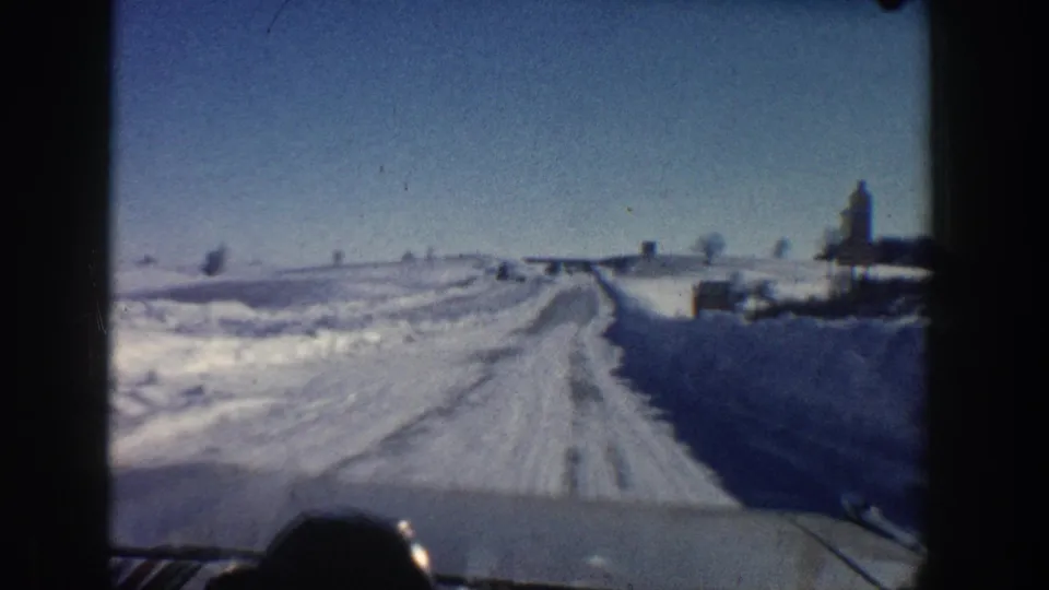 A 1961 Aspen-area windshield view shows a snowy road receding through open fields with sparse roadside buildings in the distance.
