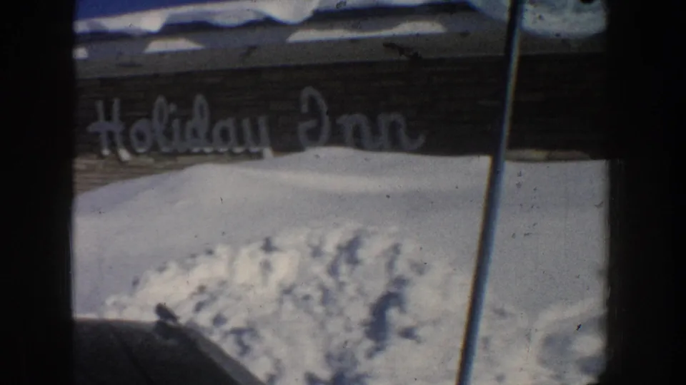 1961 Aspen home-movie frame showing a partly visible Holiday Inn sign, dark vehicle edges, and deep snow piled against a roadside building.